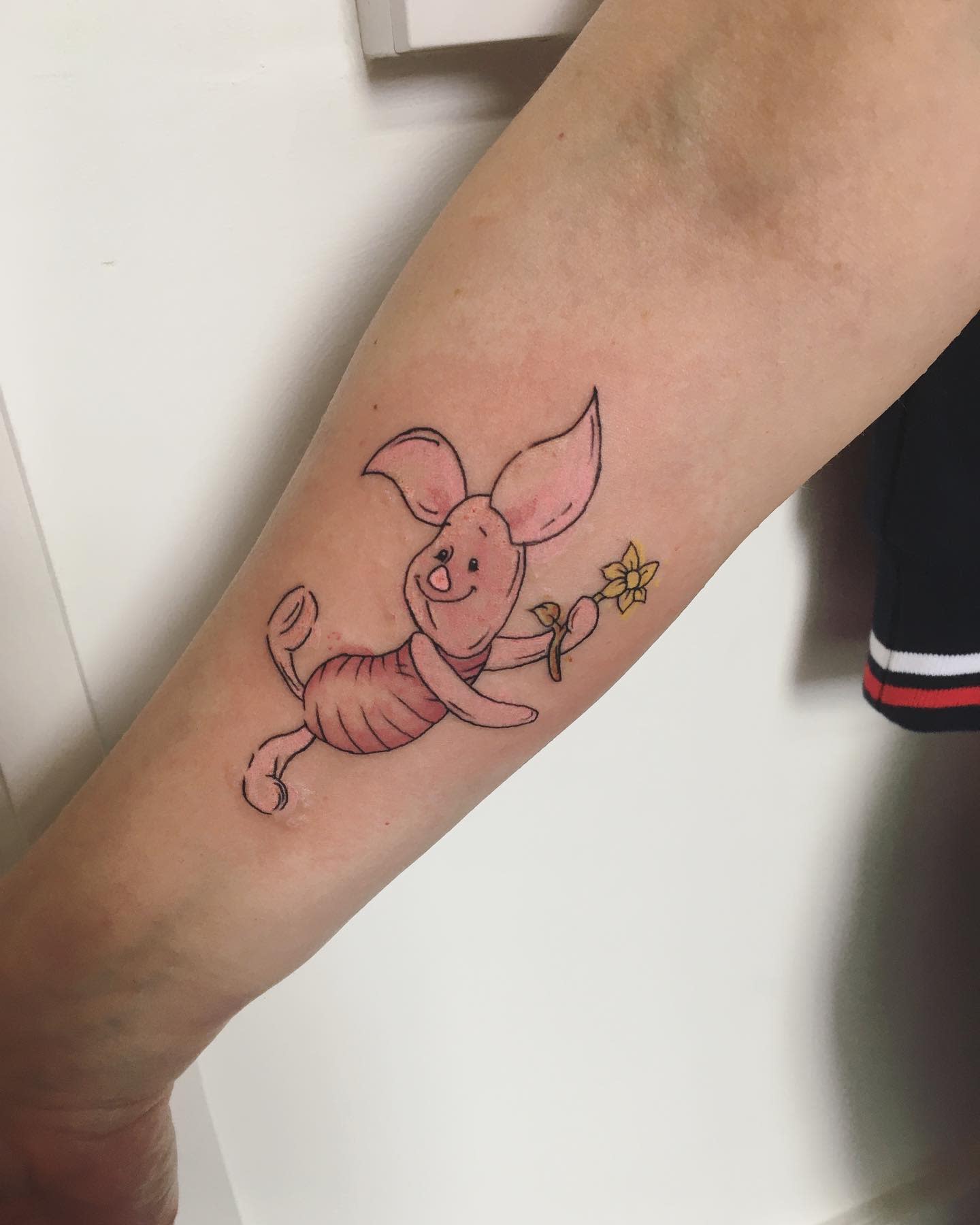 Tattoo of Piglet happily holding a small yellow flower on a person's forearm